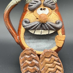 May include: A ceramic mug shaped like a character with a brown body, a large dark brown mustache, white teeth, and two eyes. The mug features a handle, a wide open mouth, and two textured brown feet extending from the bottom. The rim is speckled.