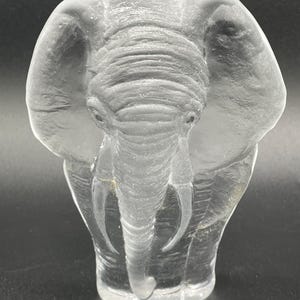 May include: A clear glass sculpture of an elephant. The elephant is standing with its trunk lowered and its ears spread out. The sculpture is detailed with lines and wrinkles to give it a realistic look.