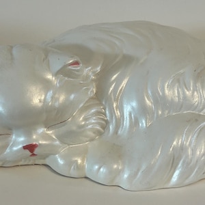 May include: A pearlescent white ceramic figurine of a sleeping cat with long, sculpted fur texture. The cat has small pink accents on the inner ears and a small red nose, depicted in a curled, resting position with eyes closed.