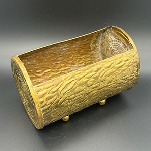 May include: A hollow, cylindrical brass container shaped like a log, featuring a textured exterior that mimics tree bark and circular ring patterns on the ends. The vessel is supported by small, rounded feet attached to the bottom surface.