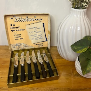 Puede incluir: Un juego de seis cuchillos de untar de metal plateado con mangos negros, presentados en una caja dorada. La tapa de la caja muestra el texto en azul y negro THE Dinkee KNIFE, An ideal spreader for BUTTER CHEESE JAM, ETC. y A PERFECT FRUIT.