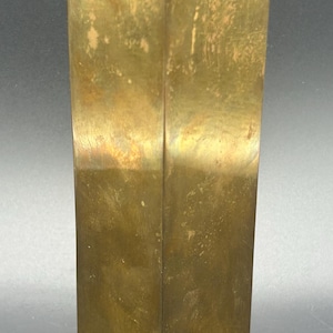 May include: A tall, square-shaped brass vase with a weathered, golden-brown patina. The object features four flat vertical sides that meet at sharp edges, leading to a circular opening at the top that reveals a metallic interior surface.