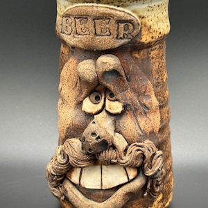 May include: A handmade ceramic beer mug with a quirky face design. The mug is brown with a tan rim and features the word "BEER" in relief. The face has a large mustache and a wide, toothy grin.