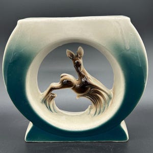 May include: A ceramic vase with a kangaroo sculpture. The vase is a rounded shape with a teal and cream color scheme. The kangaroo is brown and is leaping through a circular opening in the vase.