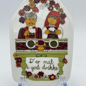 May include: A white ceramic wall plaque with a colourful illustration of a couple sitting at a table with a glass of beer. The plaque has a hole at the top for hanging. The text on the plaque reads "D'er mat i god drikke".