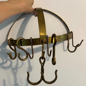 May include: A circular brass pot rack featuring a curved support frame and multiple hanging hooks. The structure includes a central twisted metal hook attachment and several S-shaped hooks suspended from the main horizontal brass band.