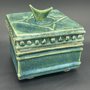 May include: A square ceramic box with a lid featuring a teal and green mottled glaze. The lid has a central triangular handle and a border of small geometric shapes including squares, circles, and triangles. The box rests on small feet.