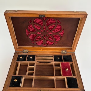 Peut inclure: Une boîte à bijoux rectangulaire en bois avec un couvercle ouvert présentant une découpe circulaire doublée de satin rouge. L'intérieur comporte plusieurs compartiments, dont quatre sections doublées de velours noir avec des boutons en laiton.