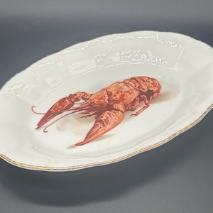 May include: An oval-shaped white ceramic serving platter featuring a detailed illustration of a reddish-brown crayfish in the center. The platter has a scalloped edge with a thin gold-colored trim along the entire outer rim of the dish.
