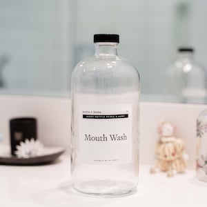 May include: Clear glass mouthwash bottle with a black cap and a white label that reads "Mouth Wash". The bottle is part of a bathroom decor set. Other decorative items are visible in the background.