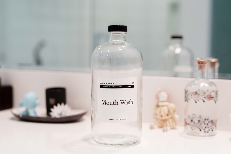 May include: Clear glass mouthwash bottle with a black cap and a white label that reads "Mouth Wash". The bottle is part of a bathroom decor set. Other decorative items are visible in the background.