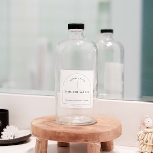 May include: A clear glass bottle with a black cap labeled "MOUTH WASH" sits on a small, round wooden stand. The label also includes the text "bottle + home" and "EVERY BOTTLE NEEDS A HOME handle with care."