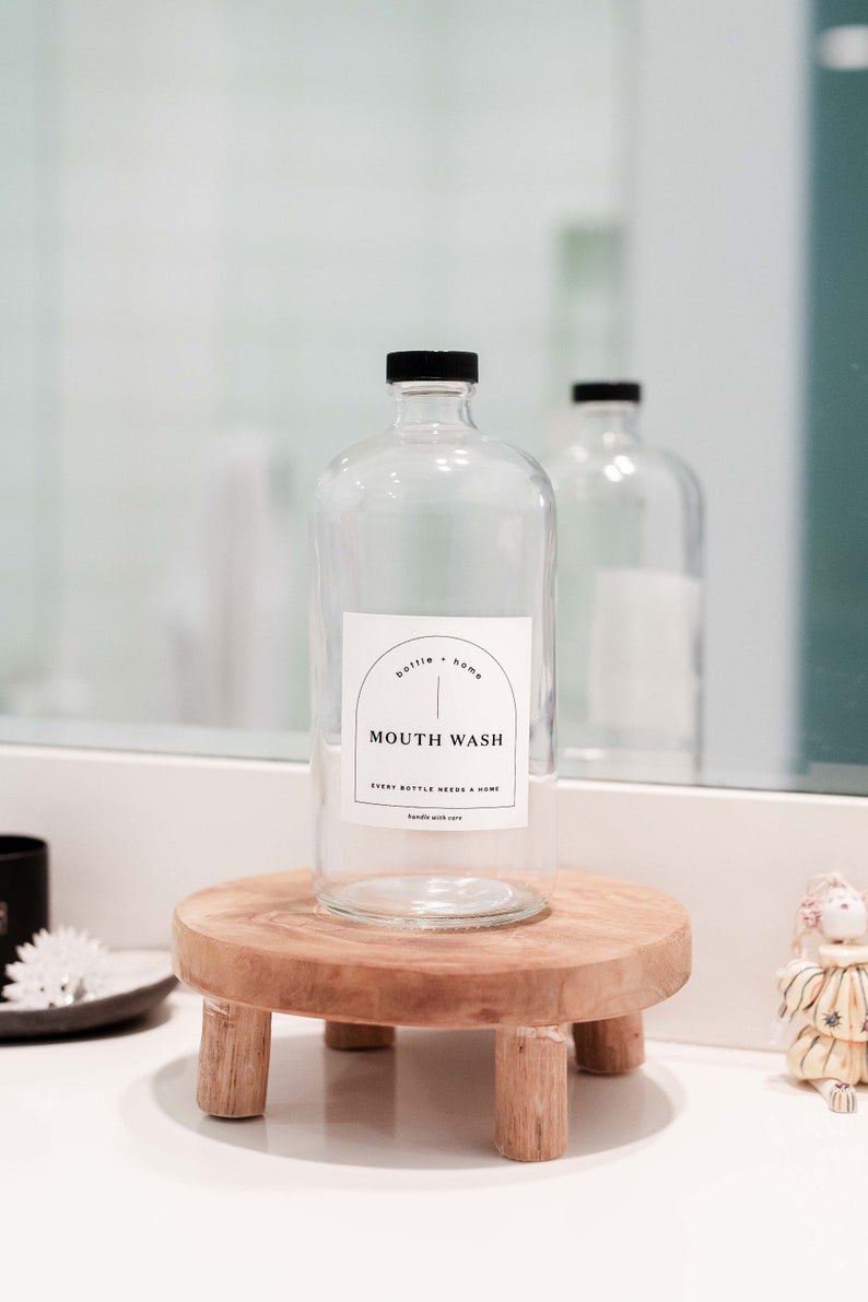 May include: A clear glass bottle with a black cap labeled "MOUTH WASH" sits on a small, round wooden stand. The label also includes the text "bottle + home" and "EVERY BOTTLE NEEDS A HOME handle with care."
