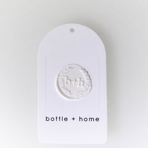 May include: White, arched tag with a debossed seal featuring the letters "b+h" within a laurel wreath. The words "bottle + home" are printed in a minimalist font below the seal. A small hole is at the top.