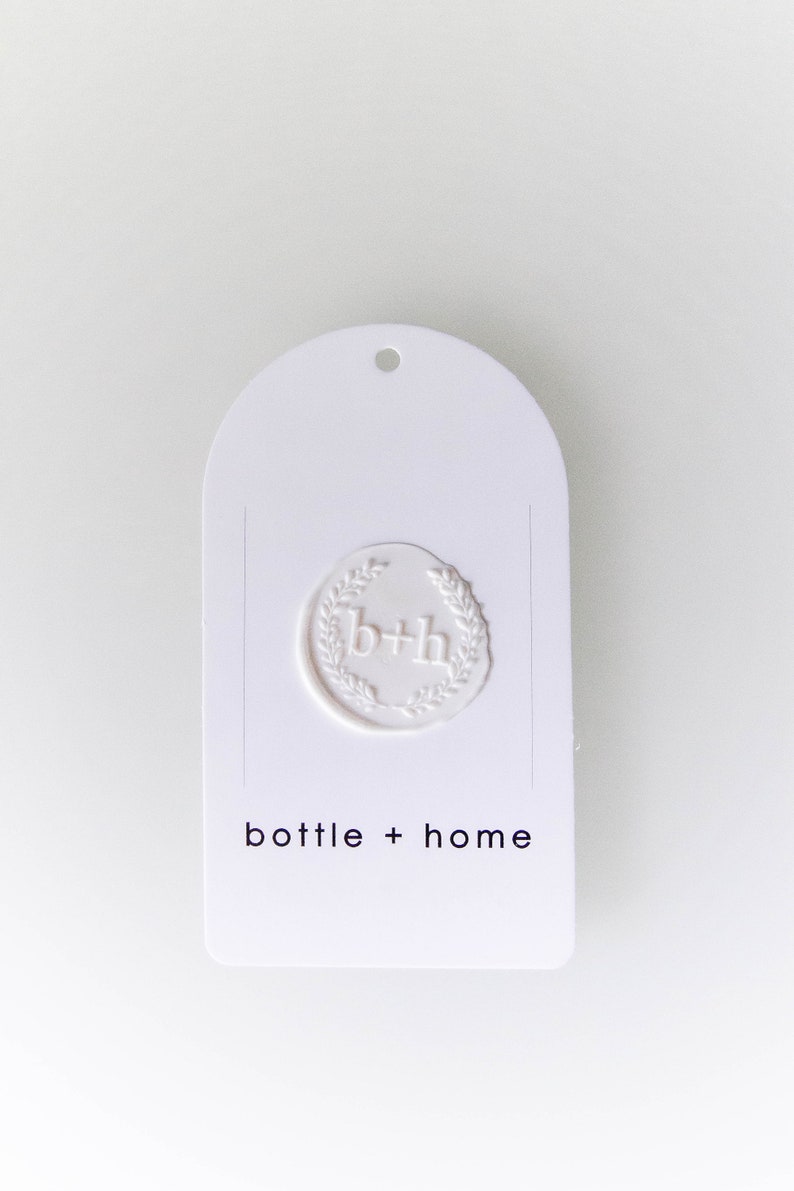 May include: White, arched tag with a debossed seal featuring the letters "b+h" within a laurel wreath. The words "bottle + home" are printed in a minimalist font below the seal. A small hole is at the top.
