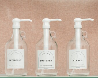 May include: Three clear glass detergent bottles with white pump dispensers. Each bottle has a white label with the words "DETERGENT", "SOFTENER", and "BLEACH". The labels also include the text "bottle + home" and "EVERY BOTTLE NEEDS A HOME".