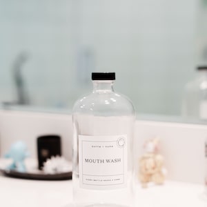 May include: Clear glass bottle with a black cap and a white label that reads "MOUTH WASH." The label also includes the text "bottle + home" and "EVERY BOTTLE NEEDS A HOME." The bottle is on a white surface.