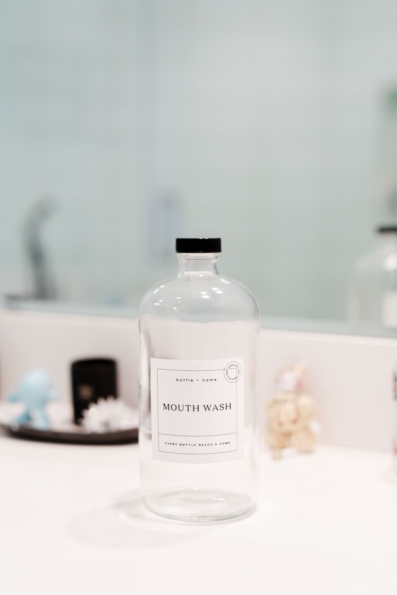 May include: Clear glass bottle with a black cap and a white label that reads "MOUTH WASH." The label also includes the text "bottle + home" and "EVERY BOTTLE NEEDS A HOME." The bottle is on a white surface.