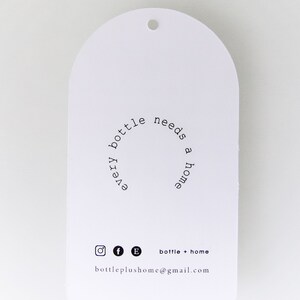 May include: White, arched tag with a small hole at the top. The tag features the text "every bottle needs a home" in a circular arrangement. Below are social media icons and the text "bottle + home" and an email address.