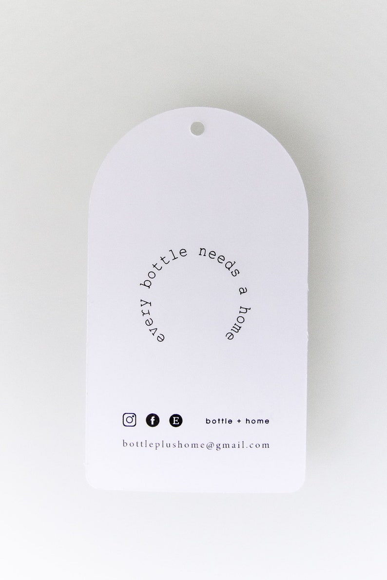 May include: White, arched tag with a small hole at the top. The tag features the text "every bottle needs a home" in a circular arrangement. Below are social media icons and the text "bottle + home" and an email address.