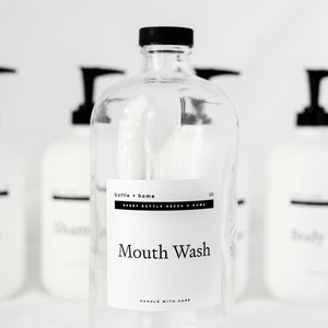 May include: Clear glass bottle with a black cap and a white label that reads "Mouth Wash." The label also includes the text "bottle + home" and "EVERY BOTTLE NEEDS A HOME." Other bottles are blurred in the background.