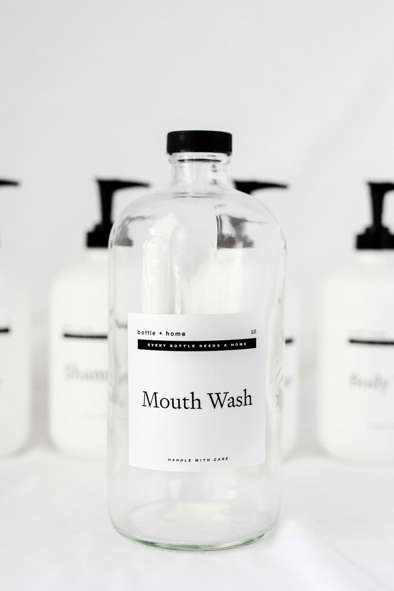 May include: Clear glass bottle with a black cap and a white label that reads "Mouth Wash." The label also includes the text "bottle + home" and "EVERY BOTTLE NEEDS A HOME." Other bottles are blurred in the background.