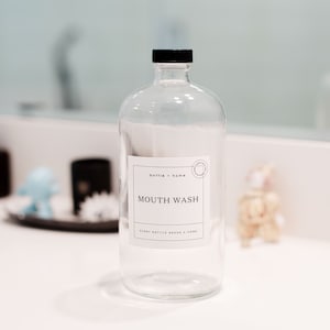 May include: A clear glass bottle with a black cap and a white label that reads "MOUTH WASH." The label also includes the text "bottle + home" and "every bottle needs a home." The bottle is on a white countertop.