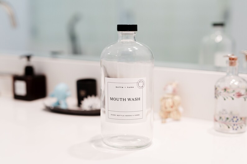 May include: A clear glass bottle with a black cap and a white label that reads "MOUTH WASH." The label also includes the text "bottle + home" and "every bottle needs a home." The bottle is on a white countertop.