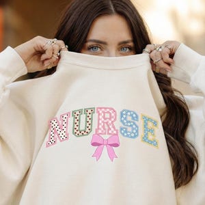 Puede incluir: Sudadera color crema con la palabra "NURSE" en letras coloridas y estampadas. Las letras tienen lunares, corazones, cuadros vichy y diseños florales. Un lazo rosa está centrado debajo de las letras. Se ven los ojos de la persona.