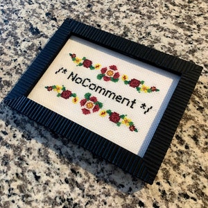 Könnte beinhalten: Ein gerahmtes Kreuzstichbild mit einem schwarzen Rahmen und weißem Stoff. Die Kreuzstich-Arbeit zeigt rote und gelbe Blumen und den Text "#NoComment#" in Schwarz.
