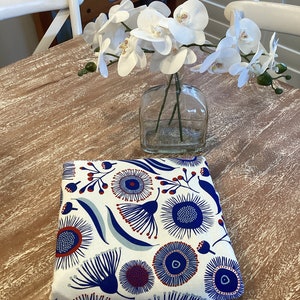 May include: A folded piece of fabric with a white background and a blue and red floral pattern. The fabric is folded on a wooden table with a clear glass vase containing white flowers.