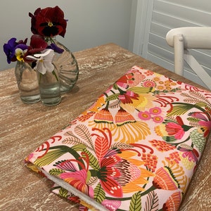 May include: A pink fabric with a colourful floral pattern featuring red, orange, yellow, and green flowers. The fabric is folded and placed on a wooden table.