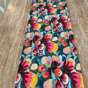 May include: A teal table runner with a colourful floral pattern. The flowers are red, pink, yellow, and orange. The runner has a stitched edge.
