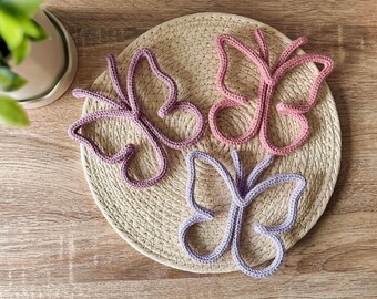 May include: Three crocheted butterflies in pink and purple colors are displayed on a woven straw mat. The butterflies are made of yarn and have a delicate, whimsical design.