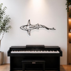May include: A black digital piano with a matching bench sits on a woven rug. A black metal shark sculpture is mounted on the white wall above the piano. A potted plant and wooden shelving unit are also visible.
