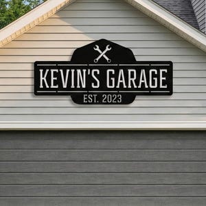 May include: A black metal sign with the text "KEVIN'S GARAGE EST. 2023" is mounted above a gray garage door. The sign features crossed wrenches and a decorative border, set against a light gray house exterior.