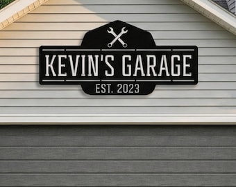 May include: A black metal sign with the text "KEVIN'S GARAGE EST. 2023" is mounted above a gray garage door. The sign features crossed wrenches and a decorative border, set against a light gray house exterior.