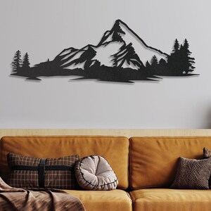 May include: Black metal wall art depicting a mountain range with snow-capped peaks and pine trees. The art is mounted on a white wall above a brown couch with pillows.