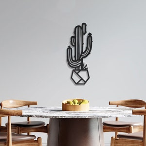 May include: Black metal wall art depicting a stylised cactus in a geometric pot. The art is displayed on a white wall above a wooden table with chairs.
