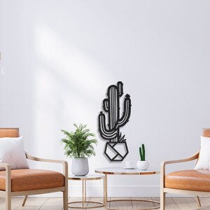 May include: Black metal wall art featuring a cactus in a geometric pot. The art is displayed in a living room setting with two armchairs and a coffee table.