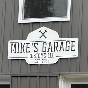 Puede incluir: Un letrero metálico rectangular blanco con una sección superior redondeada que presenta dos llaves inglesas cruzadas. El texto dice MIKE'S GARAGE, CUSTOMS LLC. y EST. 2023 en letras negras. El letrero está montado en una pared exterior gris oscuro.