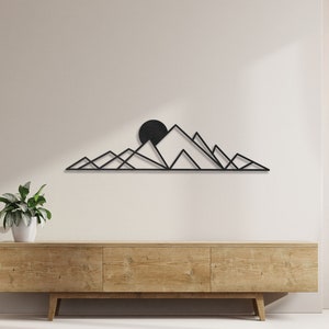 May include: Black metal wall art depicting a mountain range with a sun in the background. The art is mounted on a light wood wall shelf above a light wood cabinet.