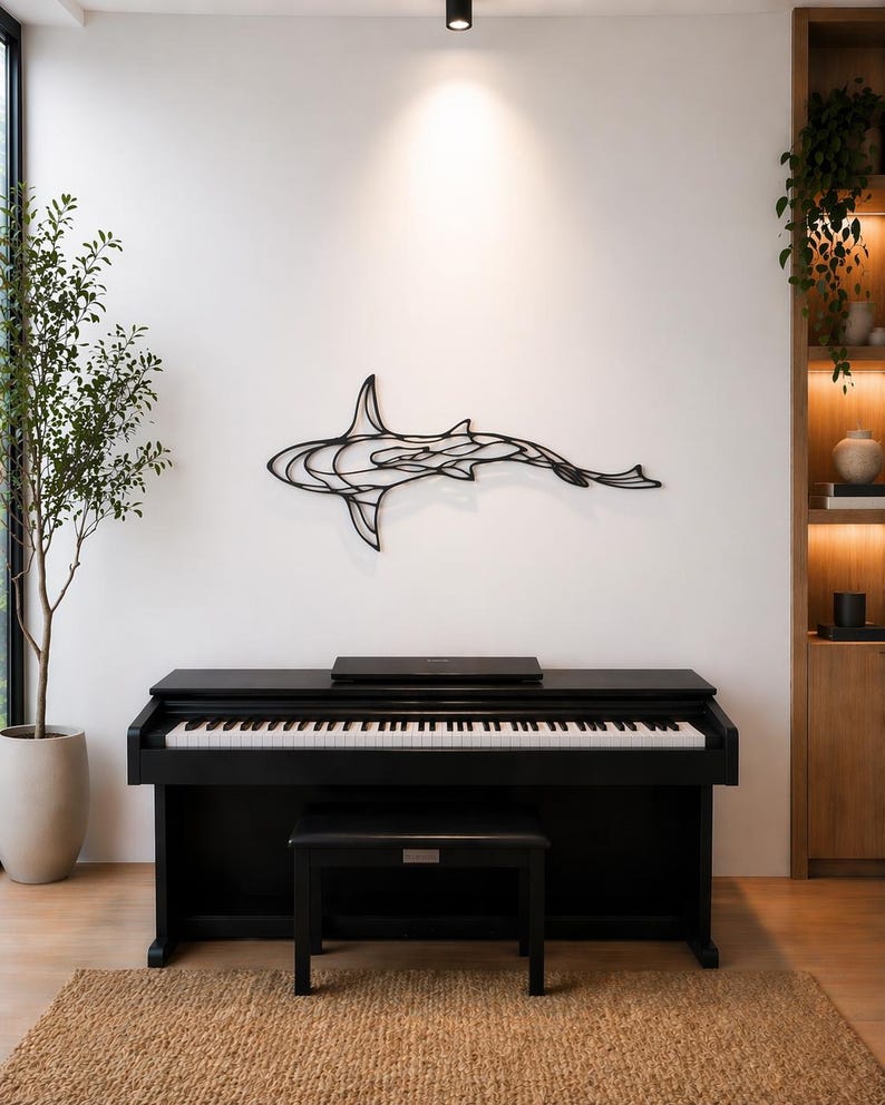 May include: A black digital piano with a matching bench sits on a woven rug. A black metal shark sculpture is mounted on the white wall above the piano. A potted plant and wooden shelving unit are also visible.