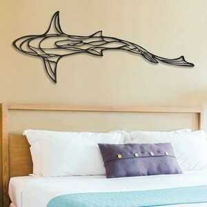 May include: A black metal shark wall art piece. The shark is depicted in a stylized, abstract design, mounted on a beige wall above a bed with white linens and a purple pillow. The room is illuminated by lamps.