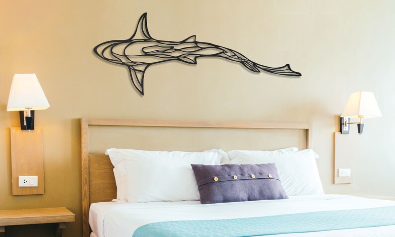 May include: A black metal shark wall art piece. The shark is depicted in a stylized, abstract design, mounted on a beige wall above a bed with white linens and a purple pillow. The room is illuminated by lamps.