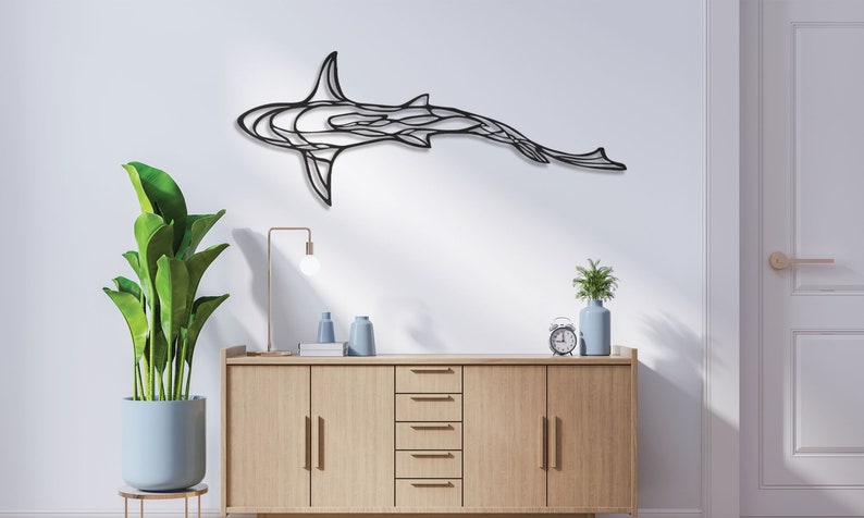 May include: A black metal shark wall art piece. The shark is depicted in a stylized, abstract design, mounted on a white wall. The room features a wooden cabinet, a potted plant, and decorative items.