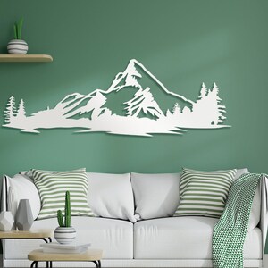 May include: White wooden wall art depicting a mountain range with trees. The art is mounted on a green wall above a white couch with striped pillows.