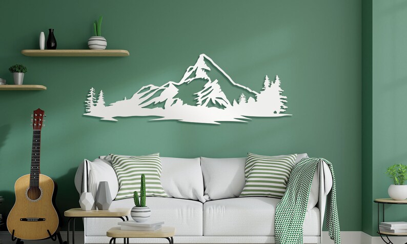 May include: White wooden wall art depicting a mountain range with trees. The art is mounted on a green wall above a white couch with striped pillows.