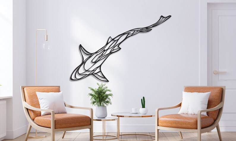 May include: A black metal shark wall art piece. The artwork is mounted on a white wall, with a modern design. The shark is depicted in a stylized, abstract form. The room features two chairs, a small table, and a plant.