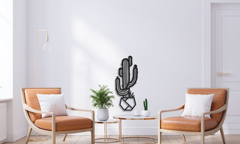 May include: Black metal wall art featuring a cactus in a geometric pot. The art is displayed in a living room setting with two armchairs and a coffee table.
